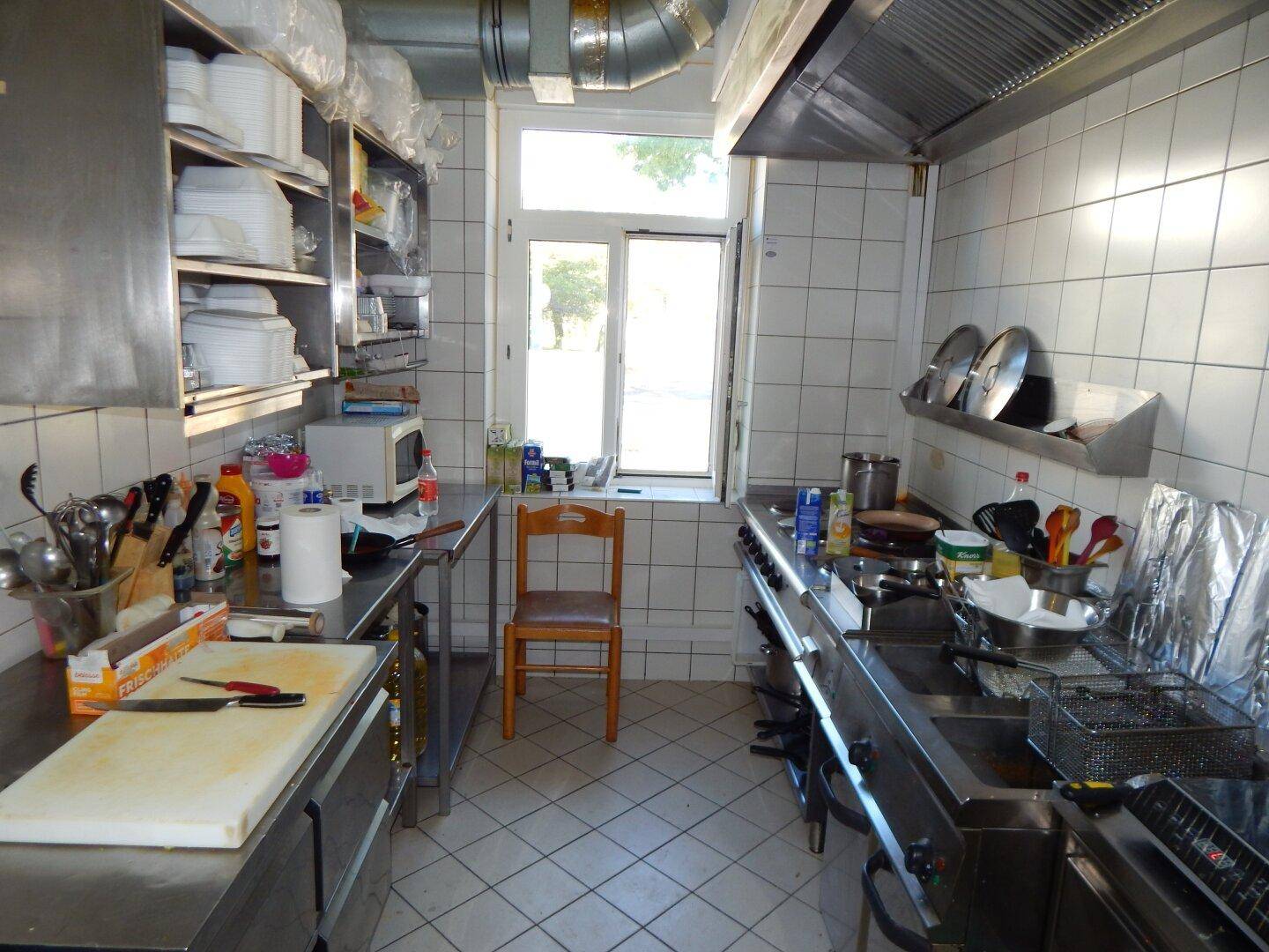 Professionelle Küche mit Edelstahlgeräten, weißen Fliesenwänden und einem Fenster für Tageslicht.