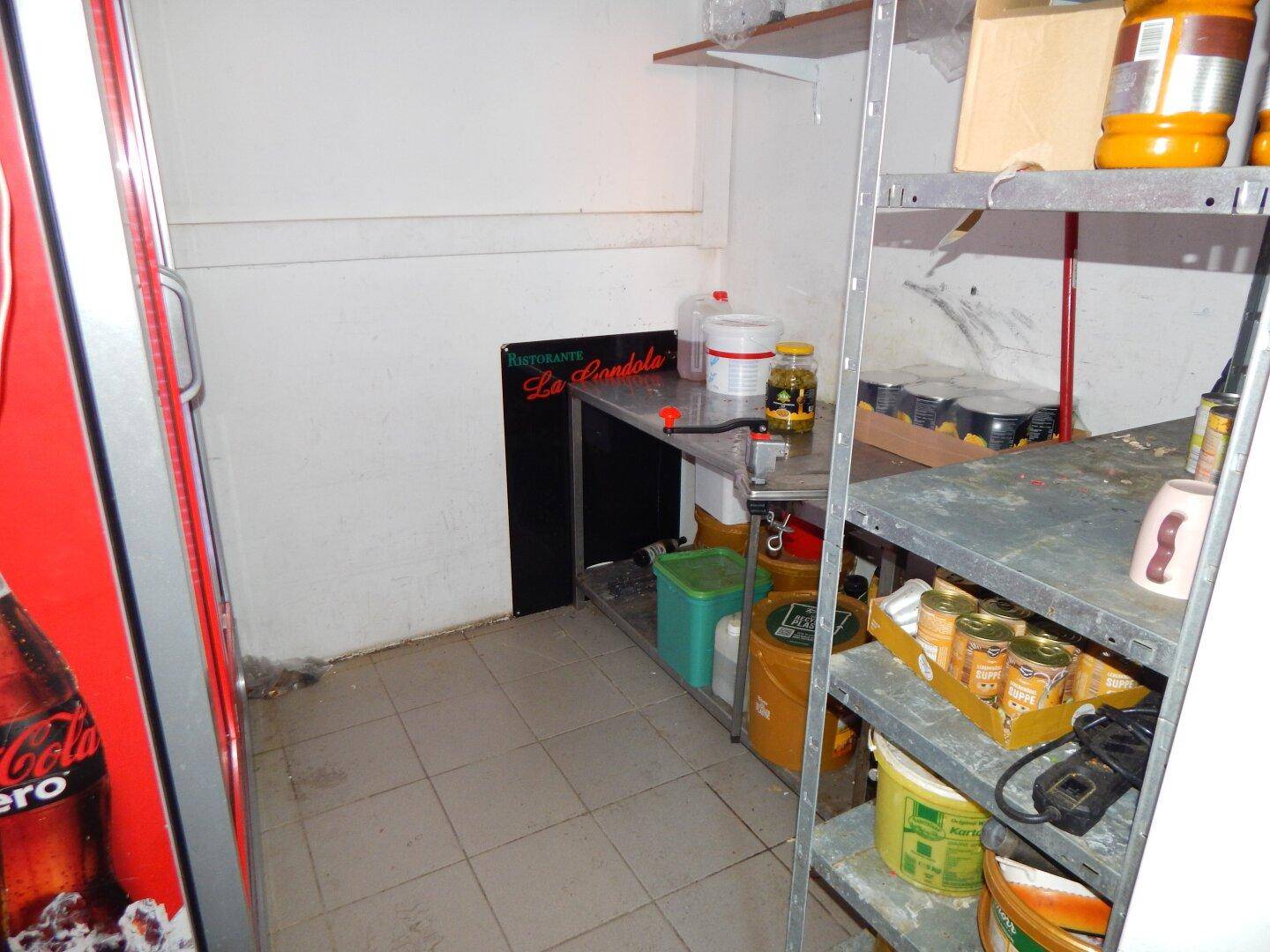 Lagerraum mit Metallregalen voller Vorräte und einem großen Kühlschrank für Lebensmittel.