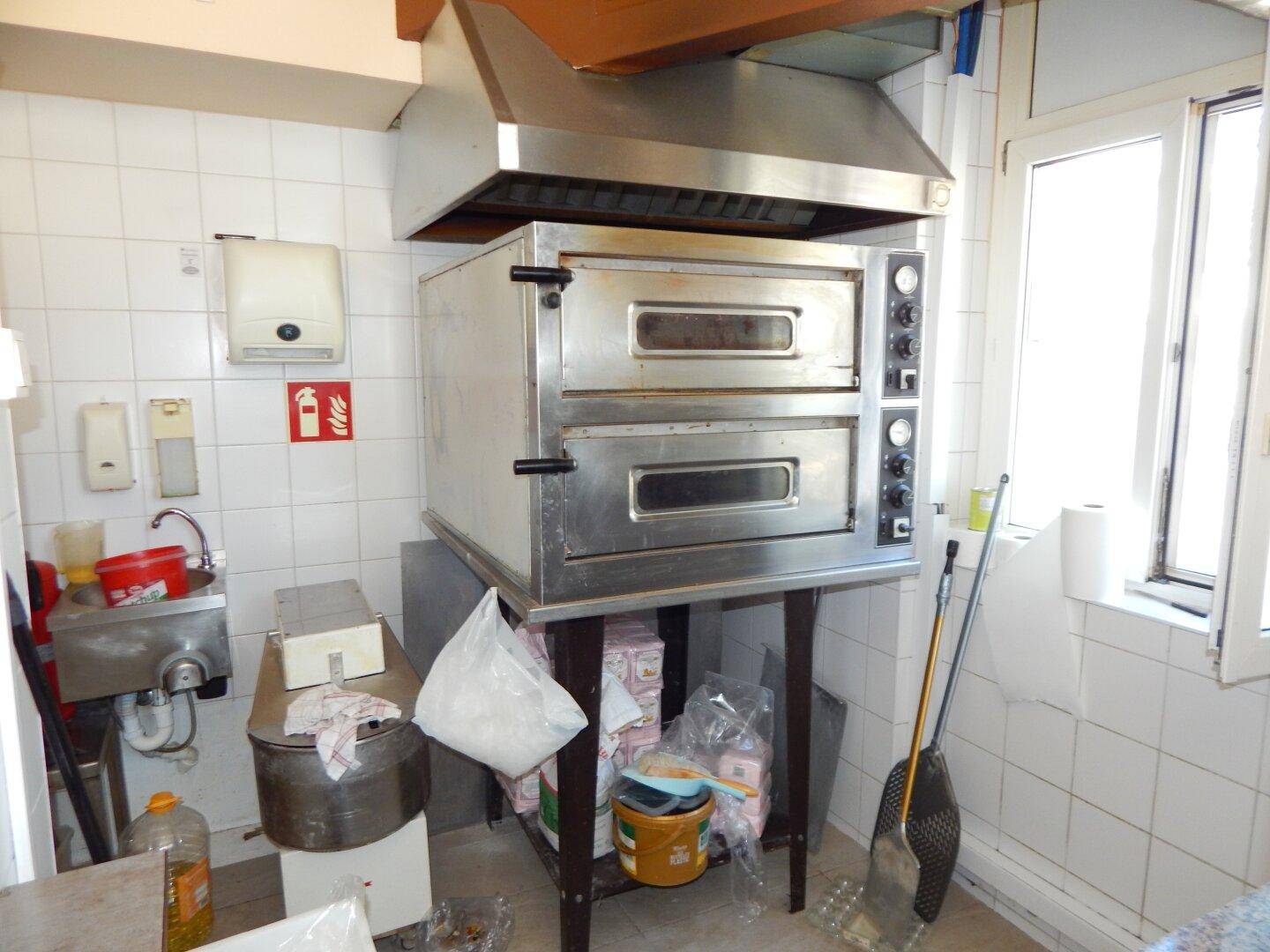 Großer Pizzaofen in einer Gewerbeküche, platziert unter einer Dunstabzugshaube neben einem Fenster.