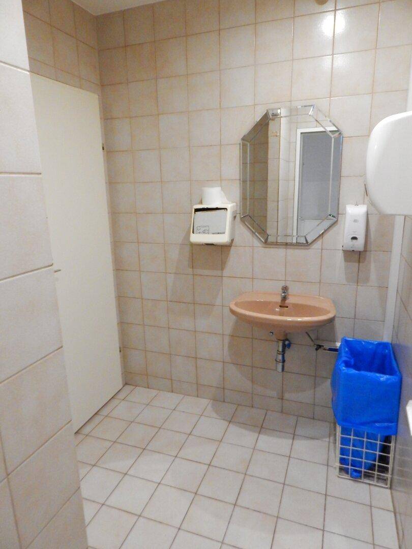 Öffentliche Toilette mit einem Waschbecken, einem achteckigen Spiegel und hellen Fliesen an Wänden und Boden.
