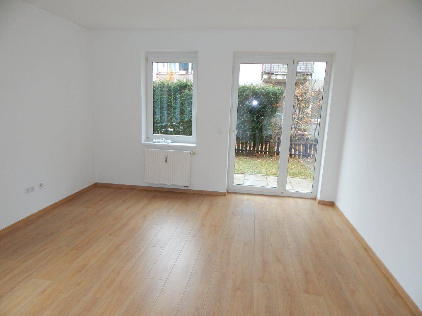 Helles, leeres Zimmer mit Laminatboden und großem Fenster mit Zugang zum Garten.