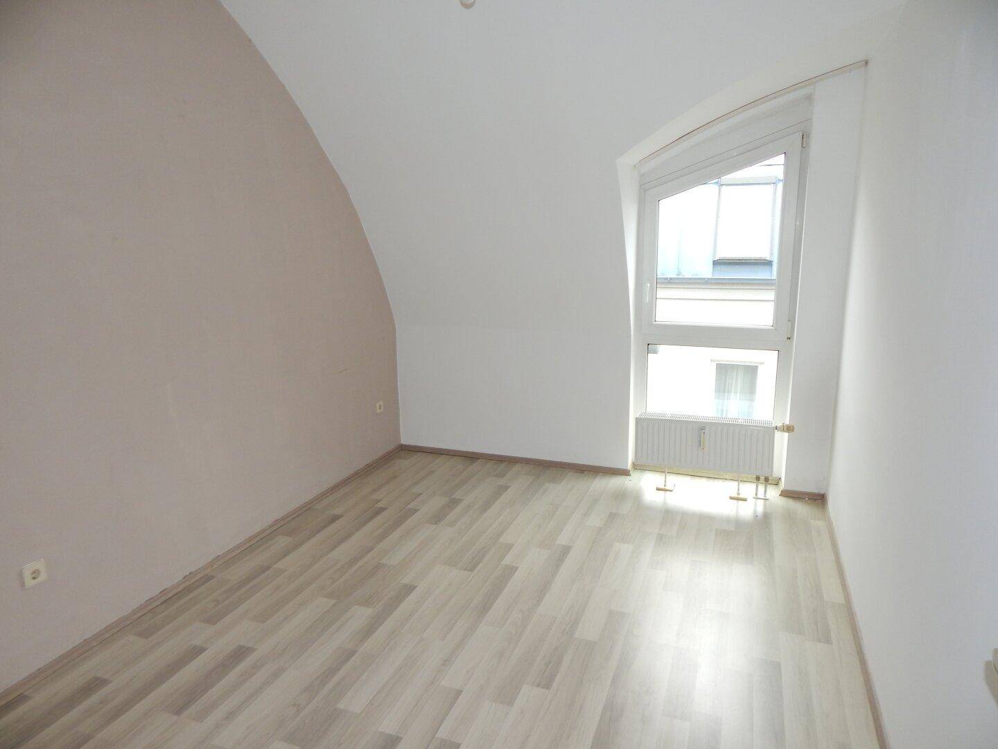 Helles Zimmer mit Dachschräge, Laminatboden und einem Fenster mit Blick nach draußen.