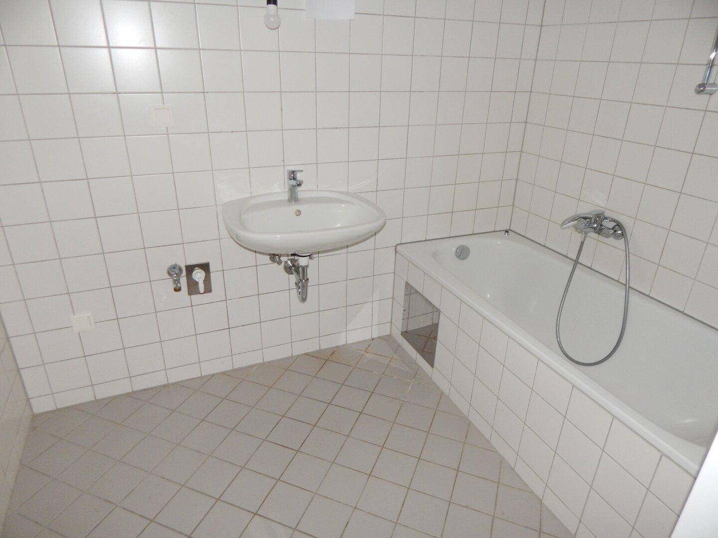 Badezimmer mit weißen Fliesen, einem Waschbecken und einer Badewanne, in einem gepflegten Zustand.