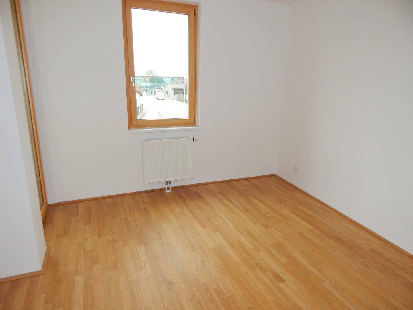 Leeres Zimmer mit Parkettboden, Heizkörper und einem Fenster, das Tageslicht spendet.
