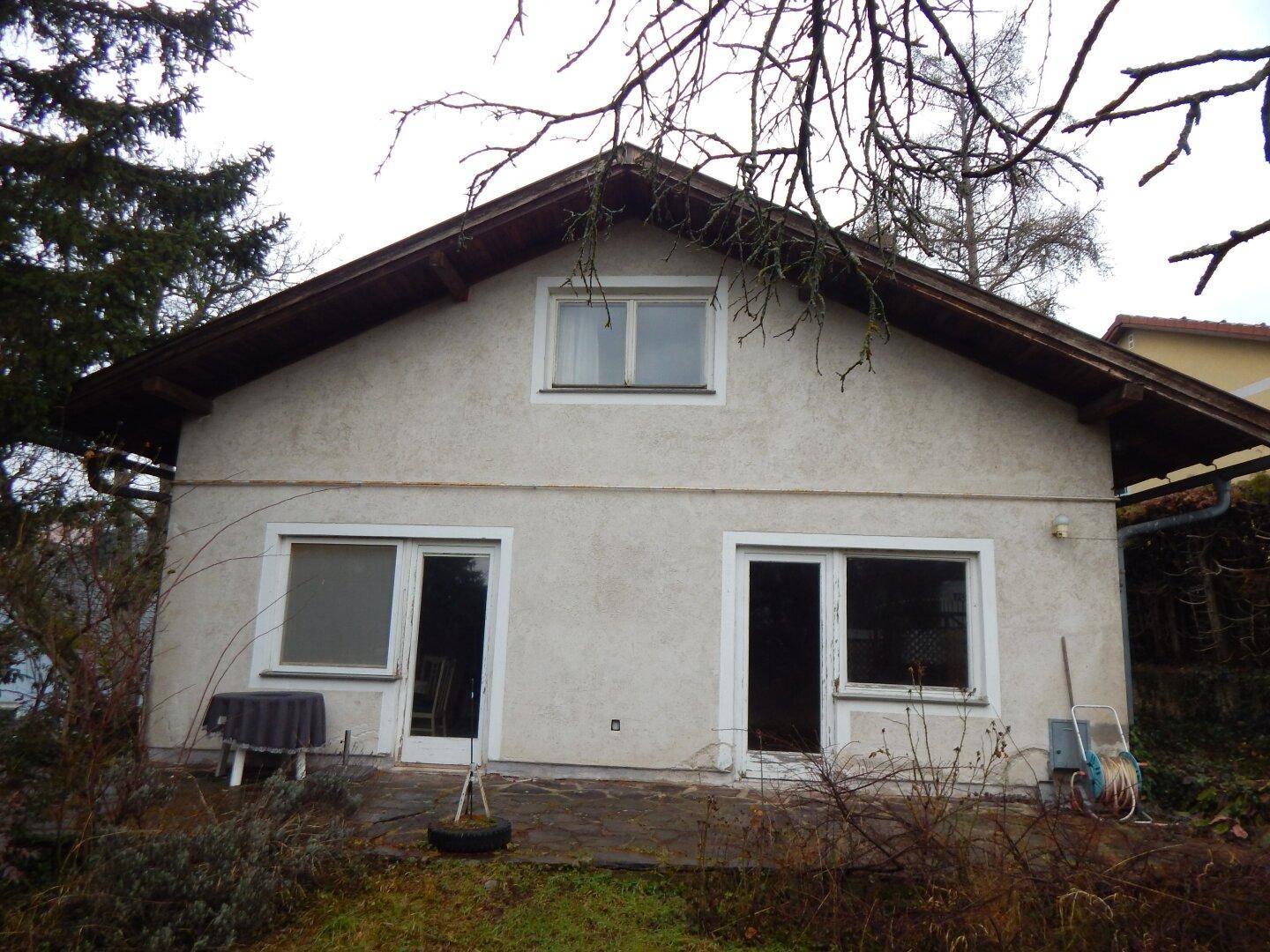 Rückseite des Hauses mit kleiner Terrasse und großen Fenstern, umgeben von Vegetation.