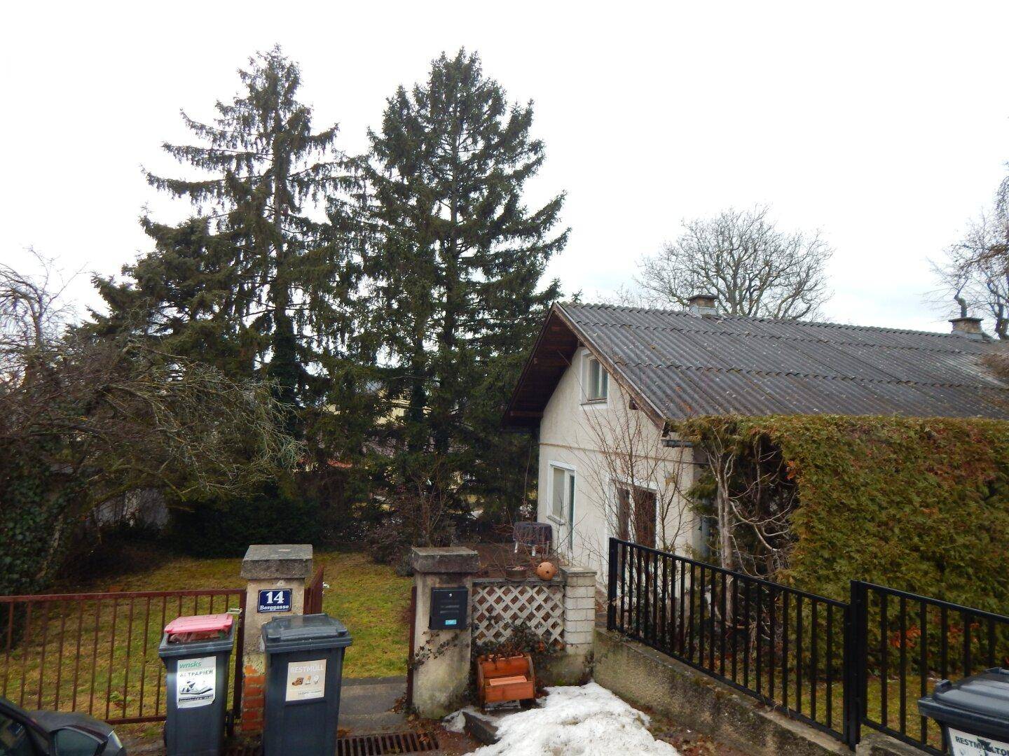 Vorderansicht des Hauses von der Straße, umgeben von Bäumen und Hecken, mit Mülltonnen.