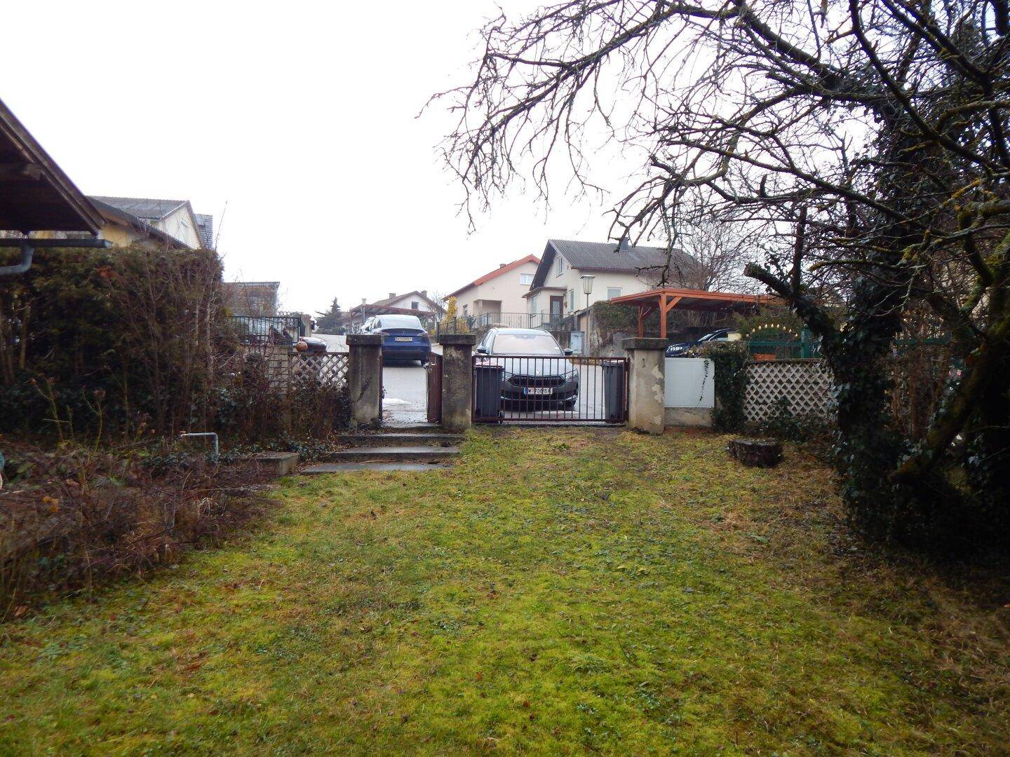 Gartenbereich mit Rasen und Blick auf die Straße und benachbarte Häuser, mit Einfahrtstor.