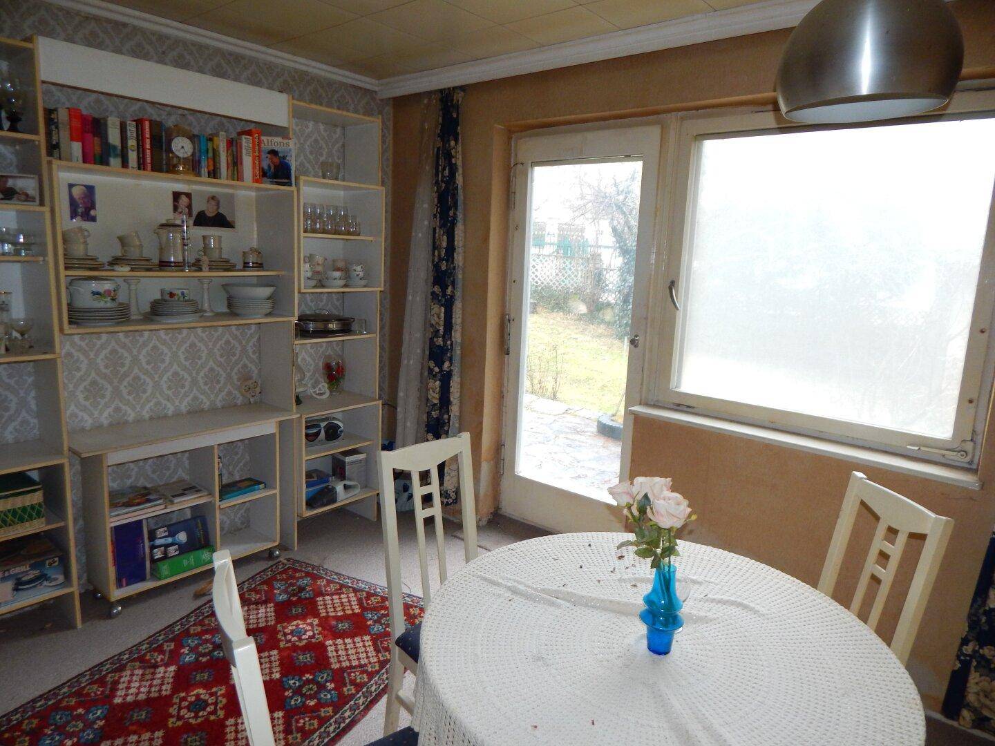 Heller Wohnbereich mit Teppichboden, Regalsystem und Blick in den Garten, mit Renovierungsbedarf.