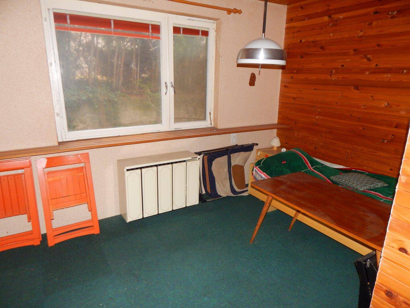 Wohnzimmer mit grünem Teppichboden, Holzvertäfelung und großem Fenster, mit Renovierungsbedarf.