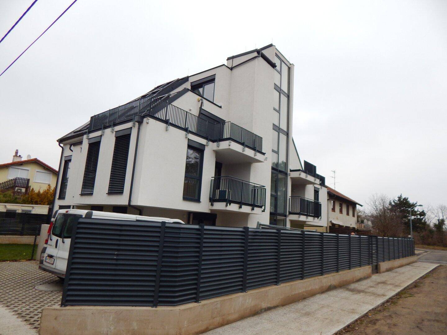 Gesamtansicht des modernen Wohngebäudes mit weißer Fassade, Balkonen und einem dunklen Lamellenzaun im Vordergrund.
