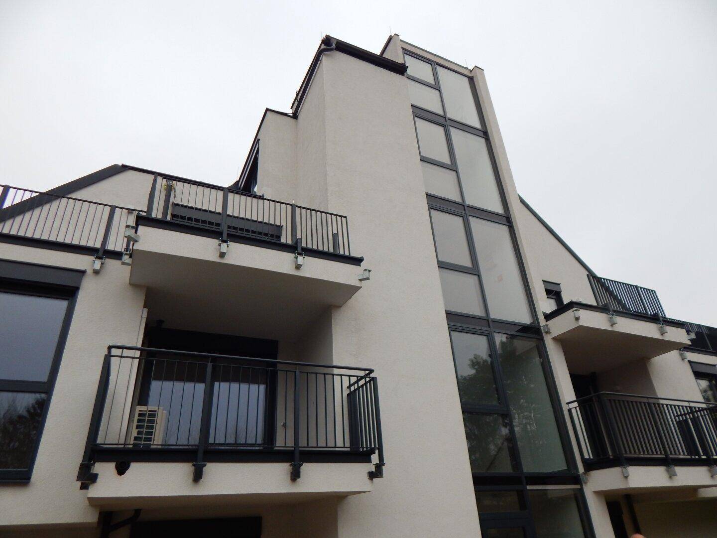 Nahaufnahme der modernen Gebäudefassade mit großen Glasflächen, Balkonen und dunklen Fensterrahmen unter bewölktem Himmel.