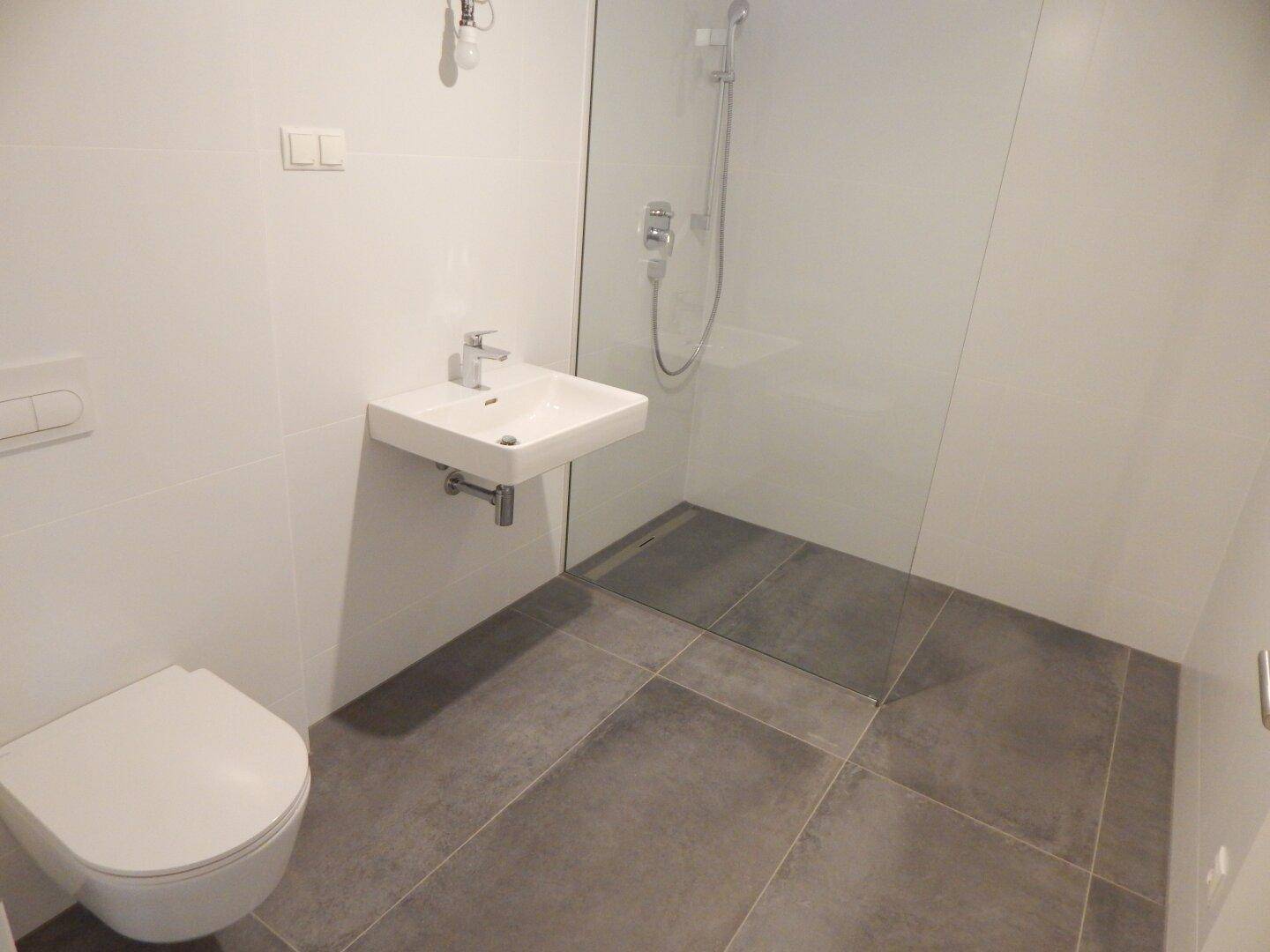 Modernes Badezimmer mit grauen Fliesen, wandhängendem WC, Waschbecken und ebenerdiger Dusche.