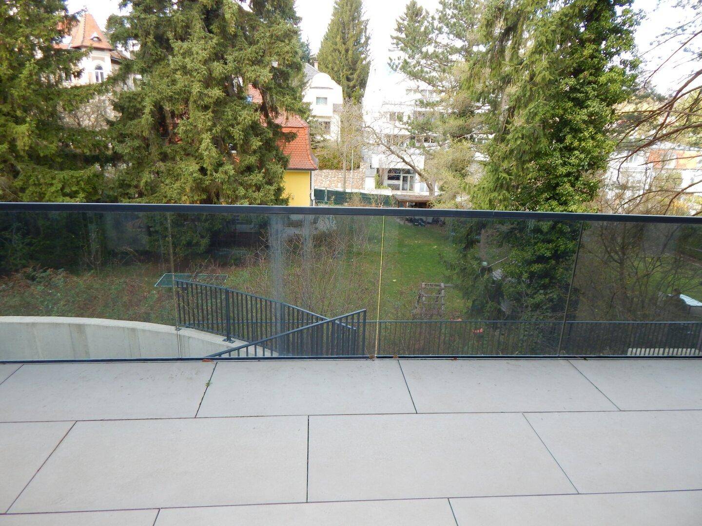 Balkon mit Glasgeländer und weitem Blick auf die grüne Umgebung und angrenzende Gärten.