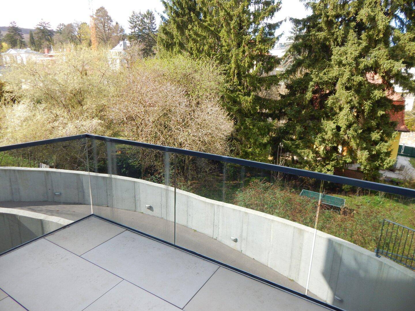 Balkon mit eleganter Glasbrüstung und Panoramablick über die grüne Landschaft und Bäume.