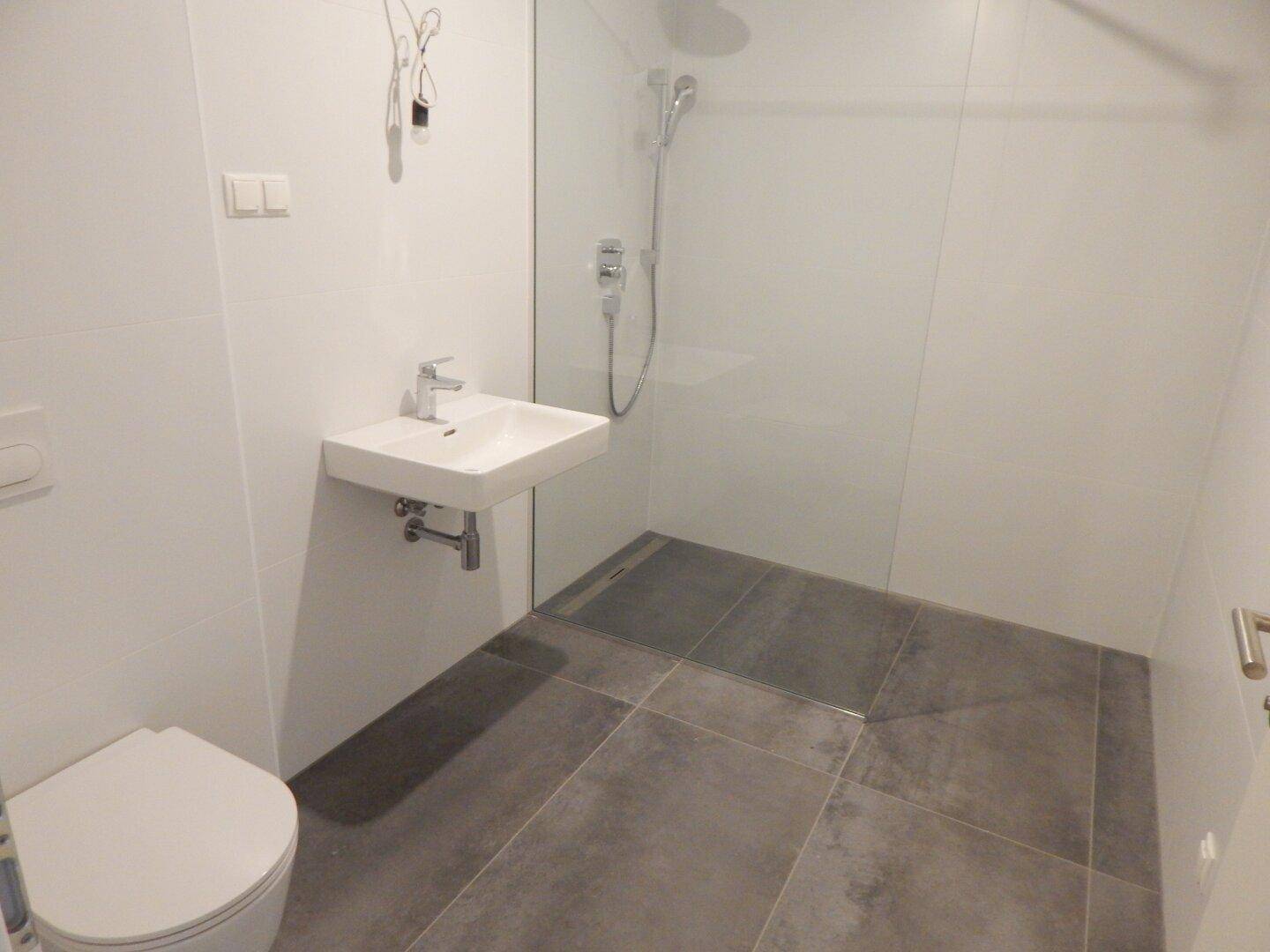 Modernes Badezimmer mit grauen Fliesen, wandhängendem WC und ebenerdiger Dusche.