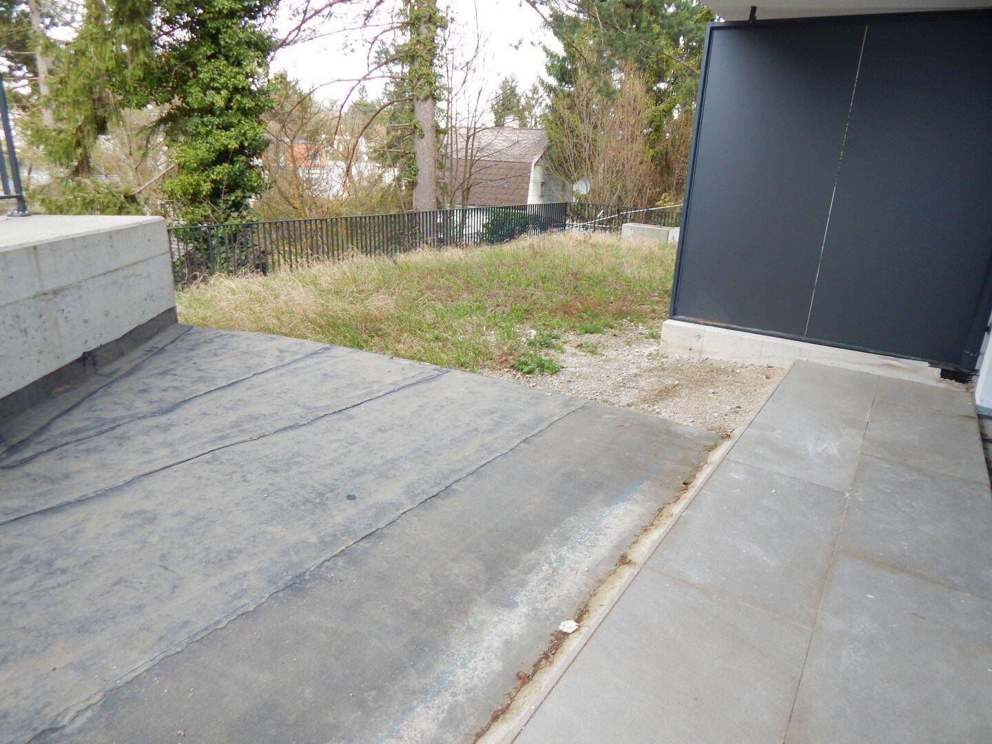 Blick auf den Außenbereich mit Betonplatten, Kies und einer angrenzenden Grünfläche.
