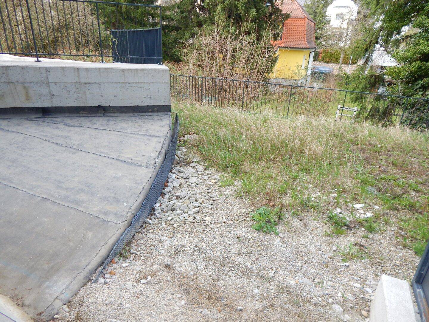 Außenbereich mit Betonfläche, Kies und Grasfläche, umgeben von einem Zaun.