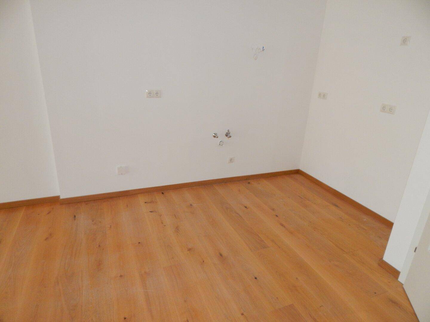 Leerer Raum mit Parkettboden und Anschlüssen für eine zukünftige Küche.