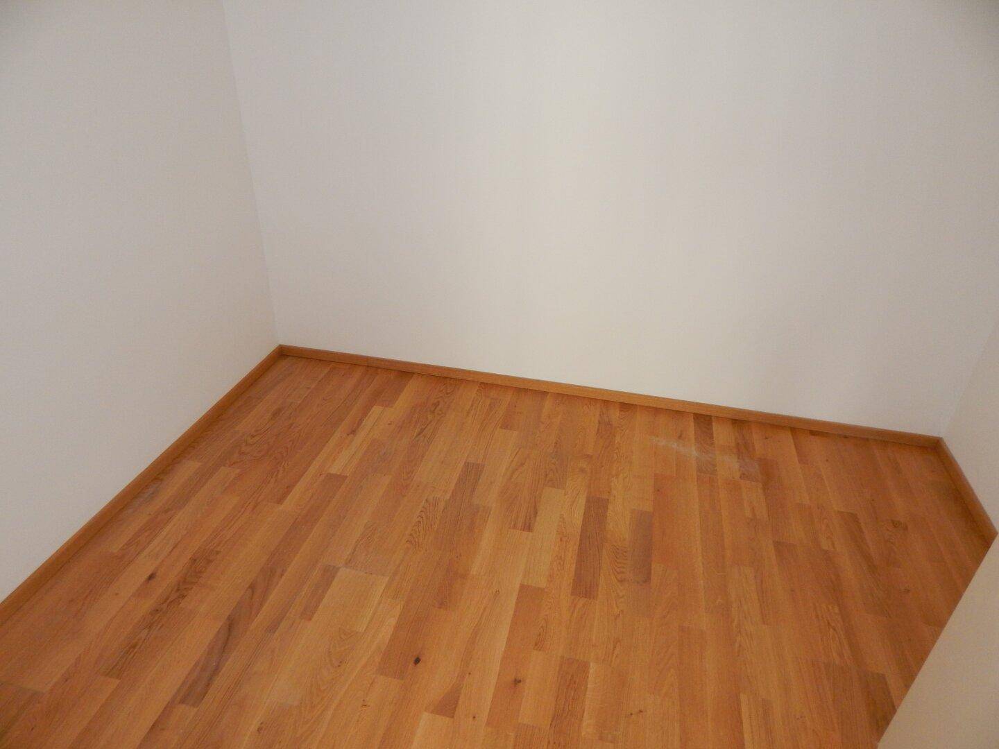 Ecke eines leeren Zimmers mit Parkettboden und weißen Wänden, bereit zur Einrichtung.