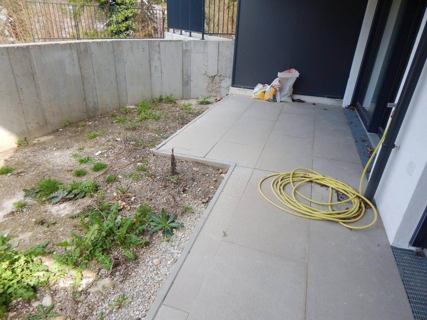 Kleine gepflasterte Terrasse mit angrenzendem, noch ungestaltetem Gartenbereich und Betonmauer.