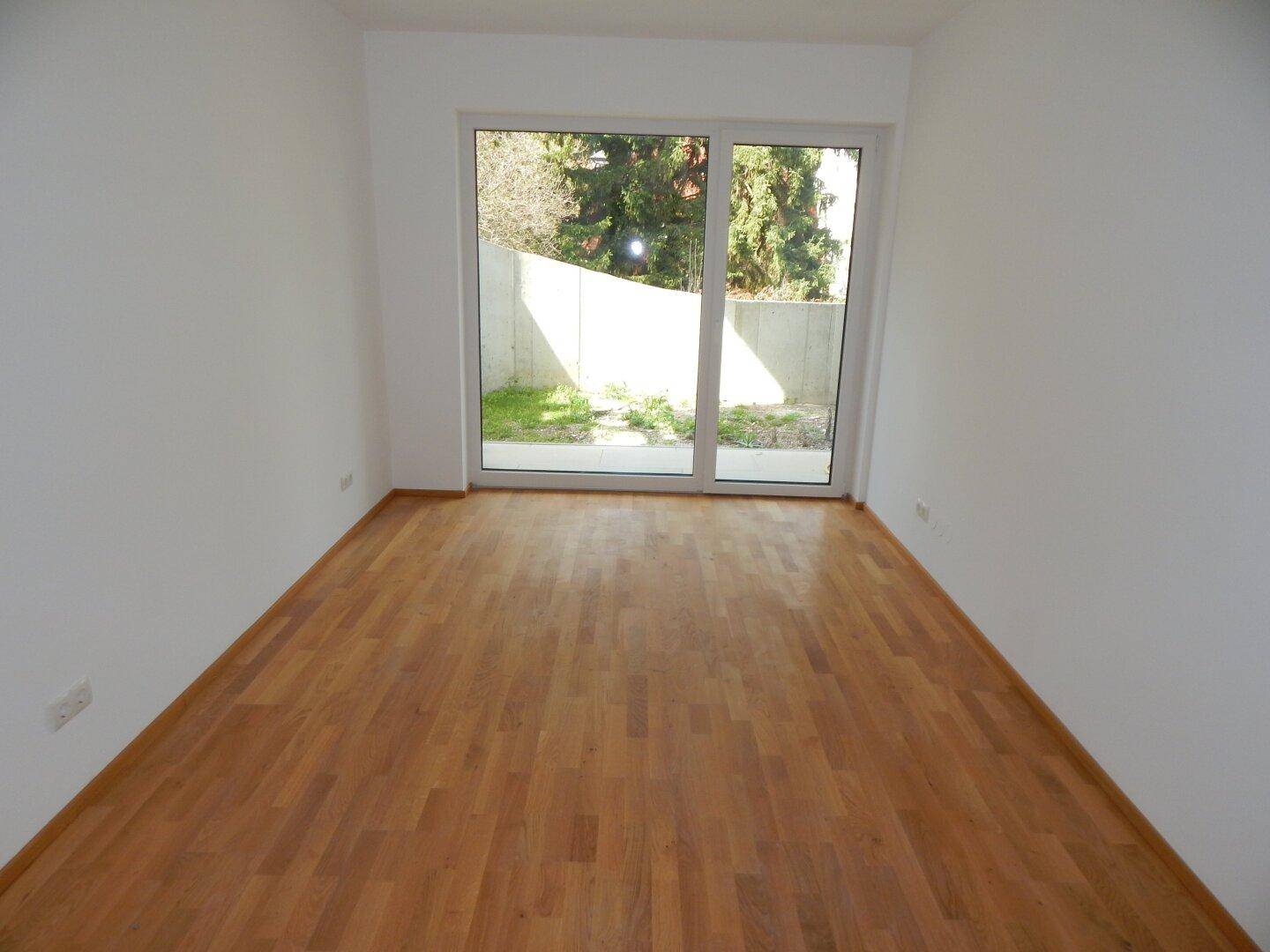 Leeres Zimmer mit Holzboden und großer Fensterfront, die viel Tageslicht hereinlässt.