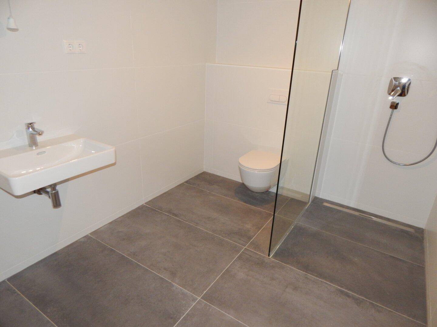 Modernes Badezimmer mit grauen Fliesen, wandhängendem WC und ebenerdiger Dusche.