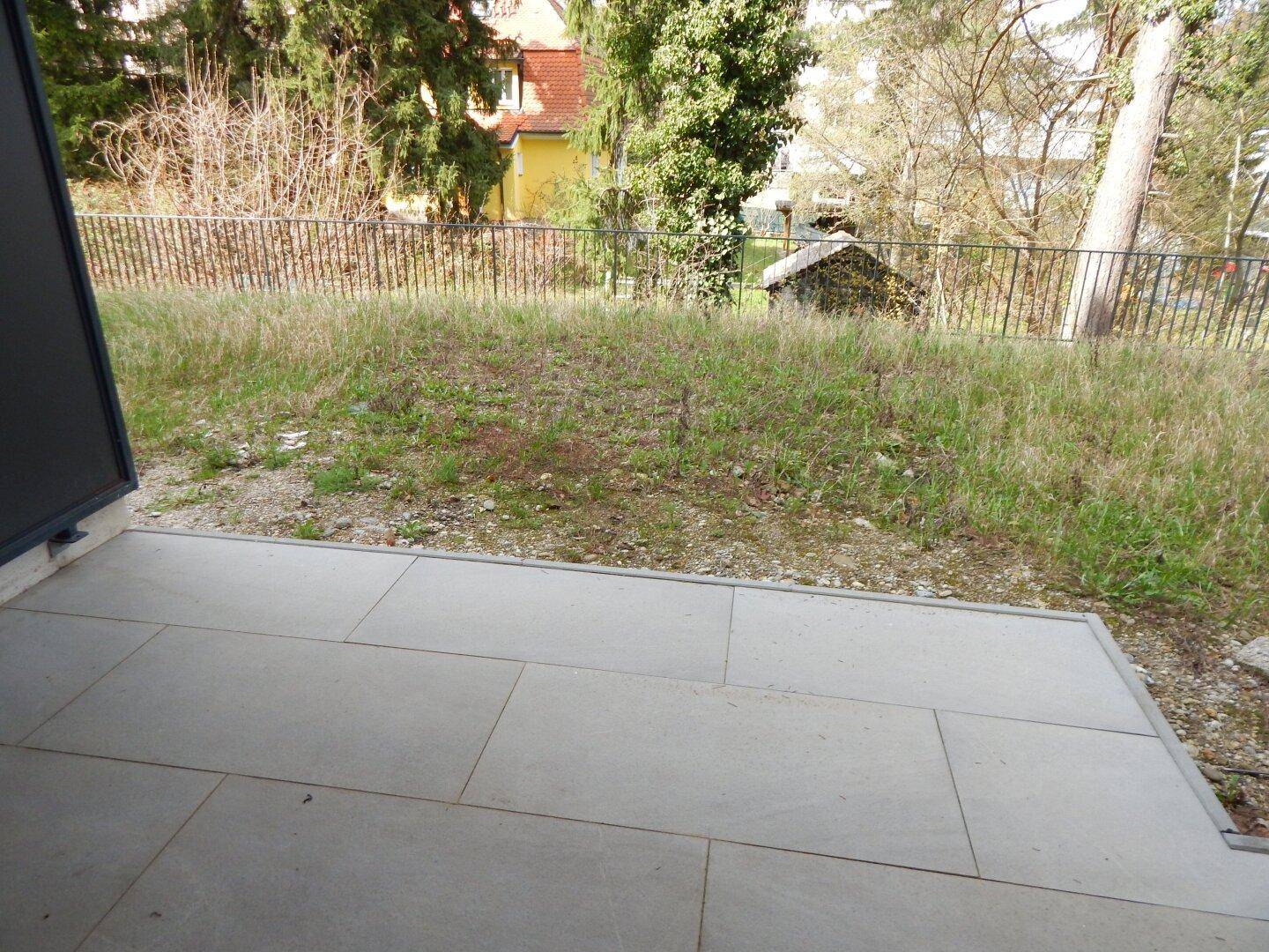 Kleine gepflasterte Terrasse mit Blick auf eine grüne Rasenfläche und ein entferntes Haus.