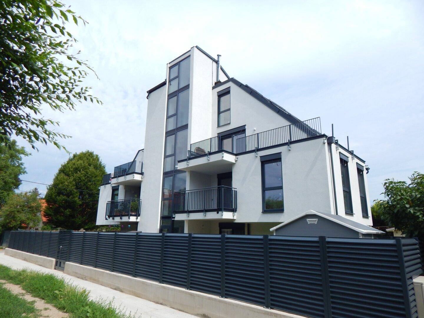 Modernes Mehrfamilienhaus mit weißer Fassade, Balkonen und dunklem Zaun in einer Wohngegend.