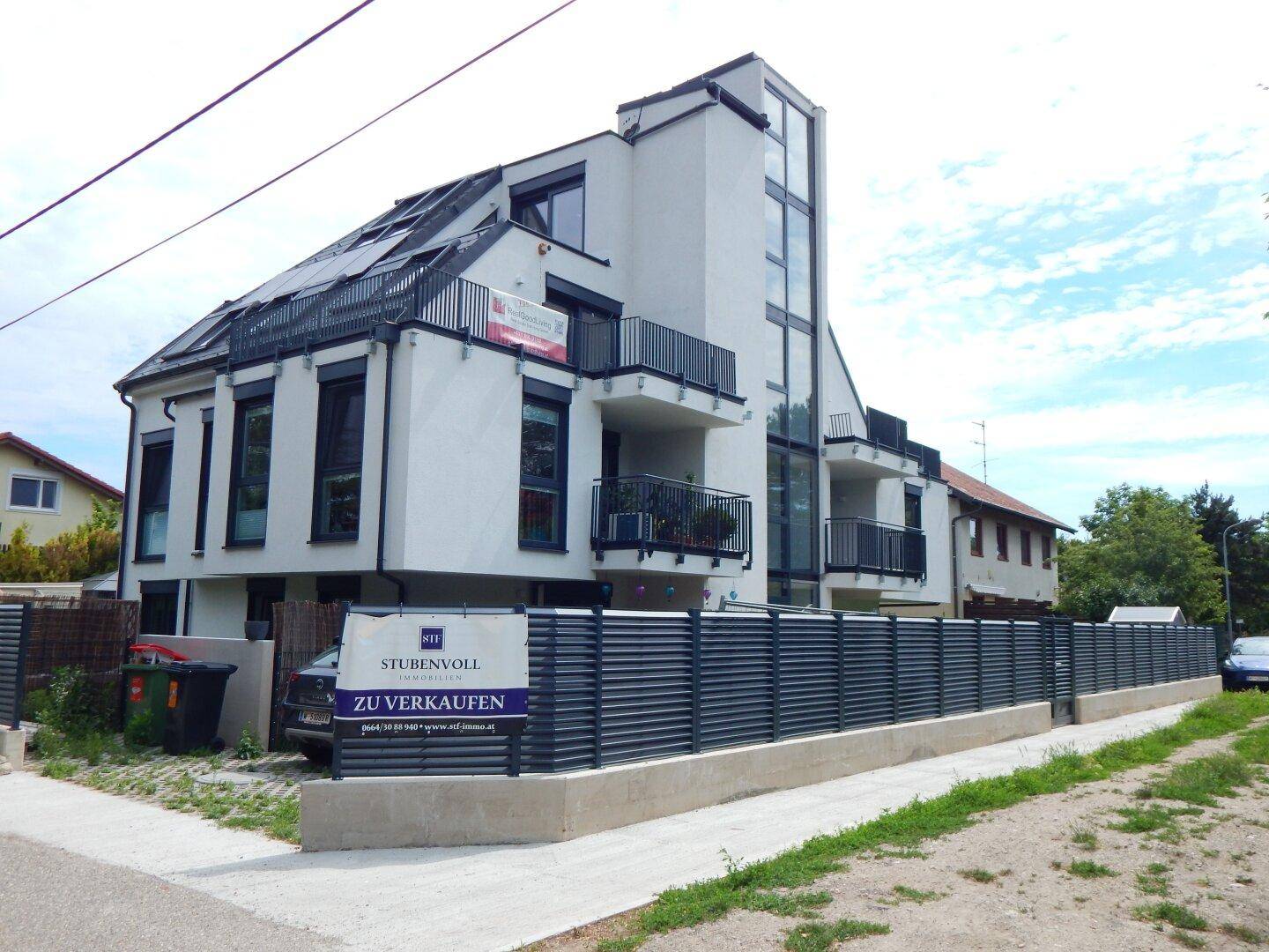 Modernes Wohngebäude mit weißer Fassade, Balkonen, Solaranlage und einem Verkaufsbanner an der Seite.