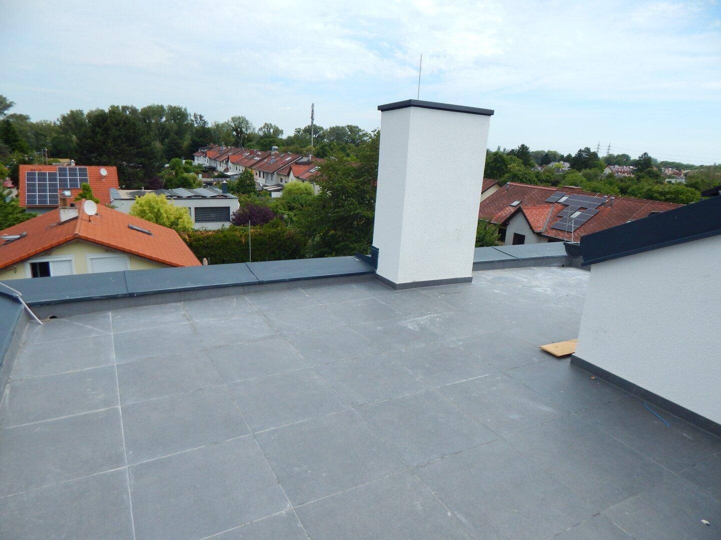 Blick von einem Flachdach mit Schornstein über die Dächer der umliegenden Häuser und viel Grün.