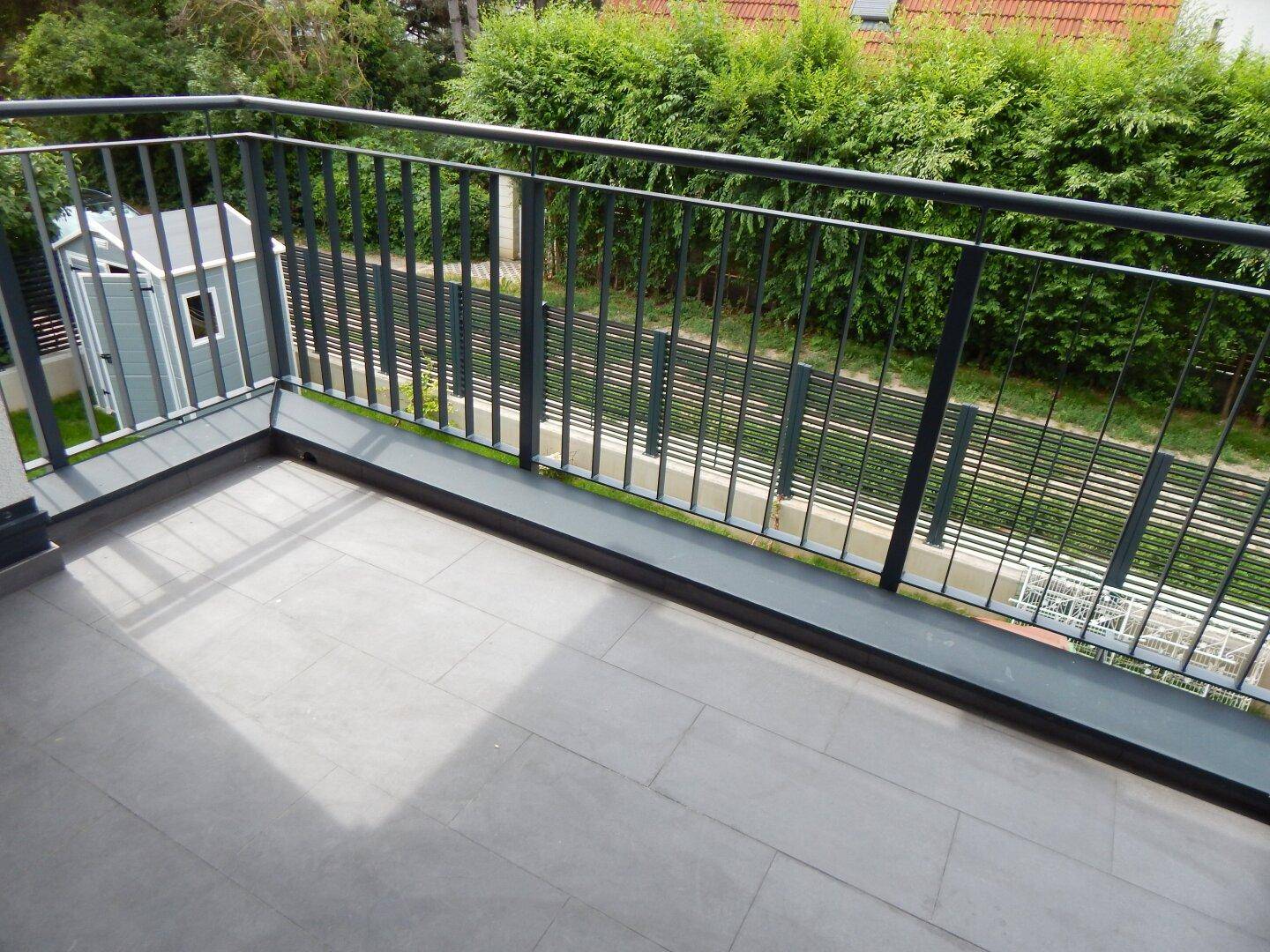 Geräumiger Balkon mit modernen Fliesen und Metallgeländer, umgeben von grünen Hecken.