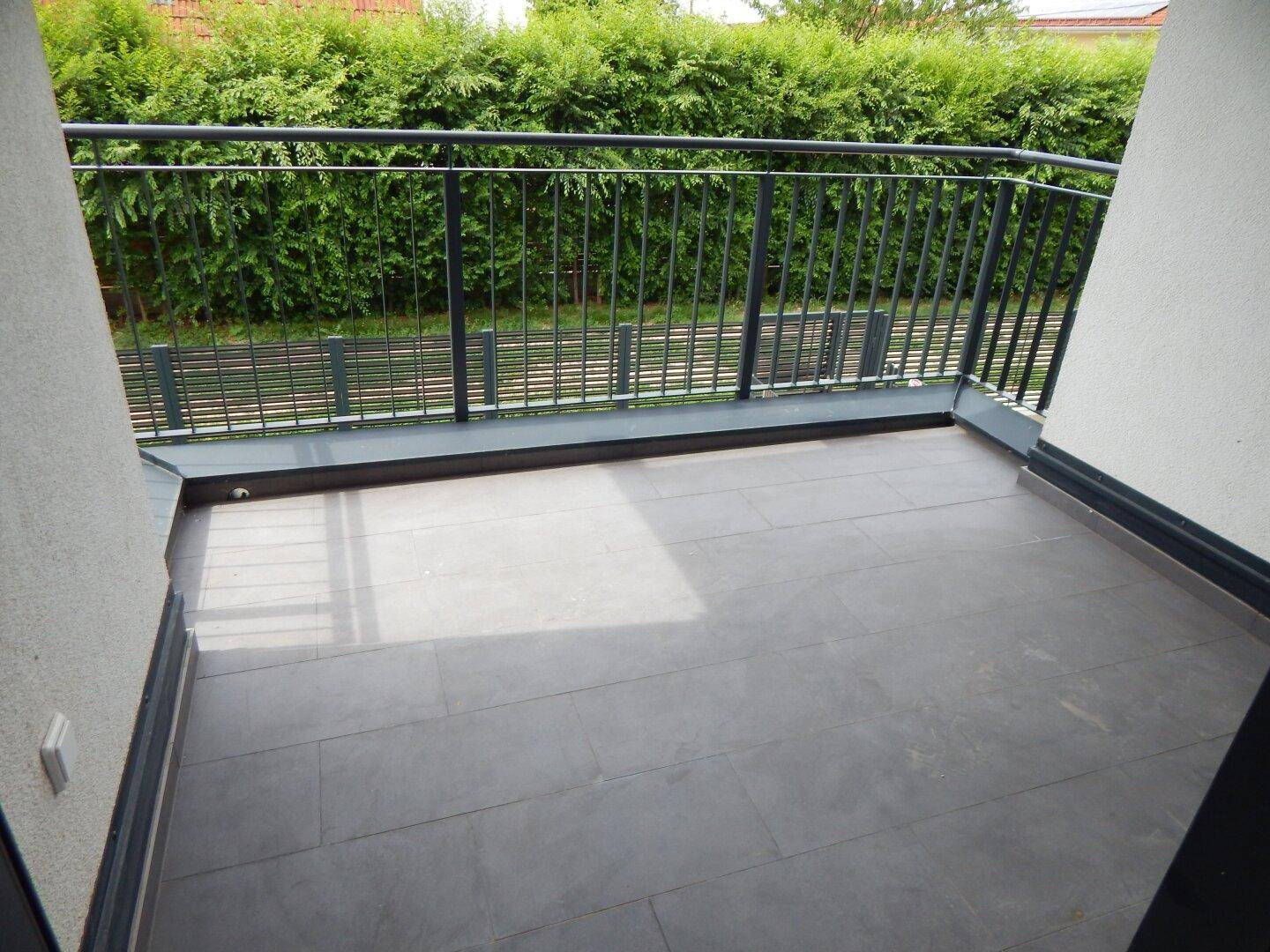 Heller Balkon mit grauen Fliesen und schwarzem Geländer, bietet einen Blick ins Grüne.