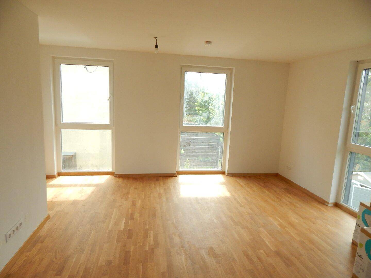 Lichtdurchfluteter Wohnraum mit hellem Parkettboden und großen Fenstern, die viel Tageslicht hereinlassen.
