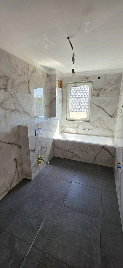 Modernes Badezimmer im Bau mit Marmorfliesen an den Wänden, dunklen Bodenfliesen und einer eingebauten Badewanne.