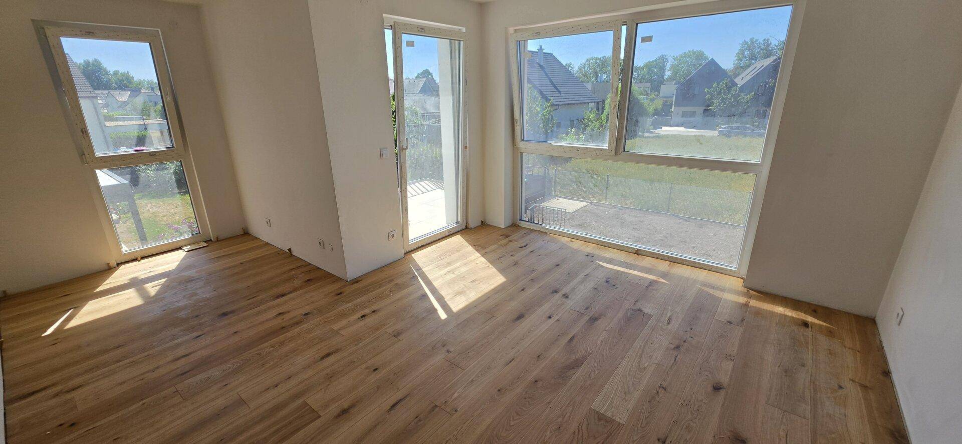 Helles Zimmer mit Holzboden und großen Fenstern, die viel Tageslicht hereinlassen und Zugang nach draußen bieten.