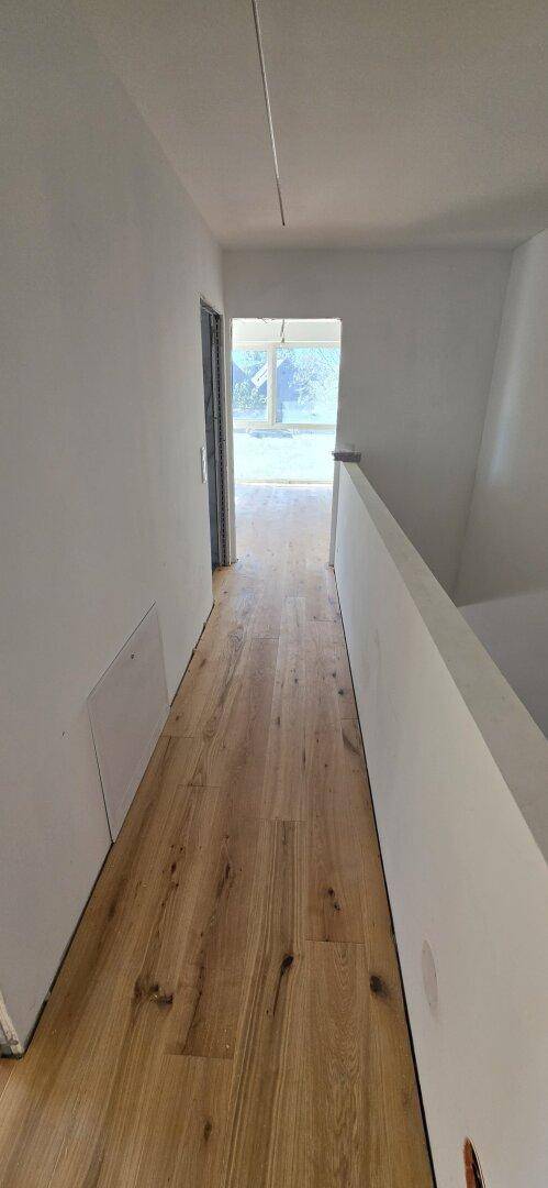 Heller Flur mit Holzboden und weißen Wänden, der zu einem weiteren Raum führt und eine offene Treppe andeutet.