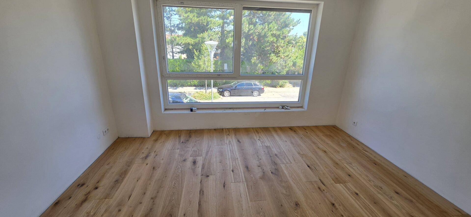 Leeres Zimmer mit hellem Holzboden und einem großen Fenster mit Blick ins Grüne und auf die Straße.