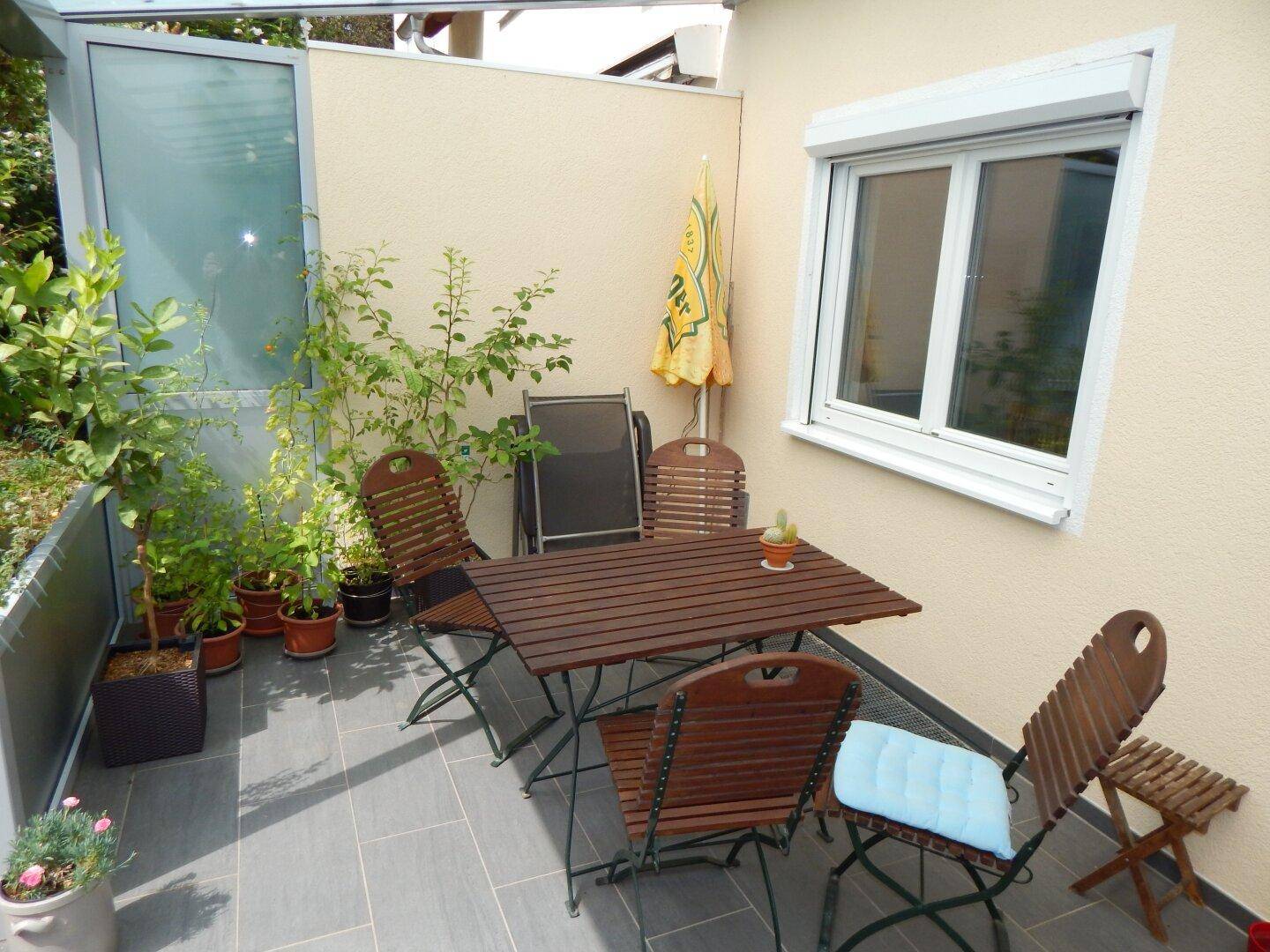 Kleine, geflieste Terrasse mit Holztisch und Stühlen, umgeben von Topfpflanzen.