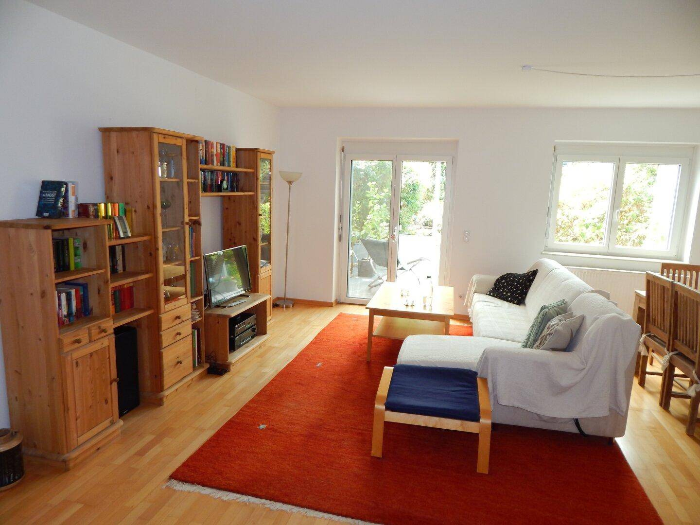 Helles Wohnzimmer mit Holzregalen, beigem Sofa, rotem Teppich und Zugang zur Terrasse.