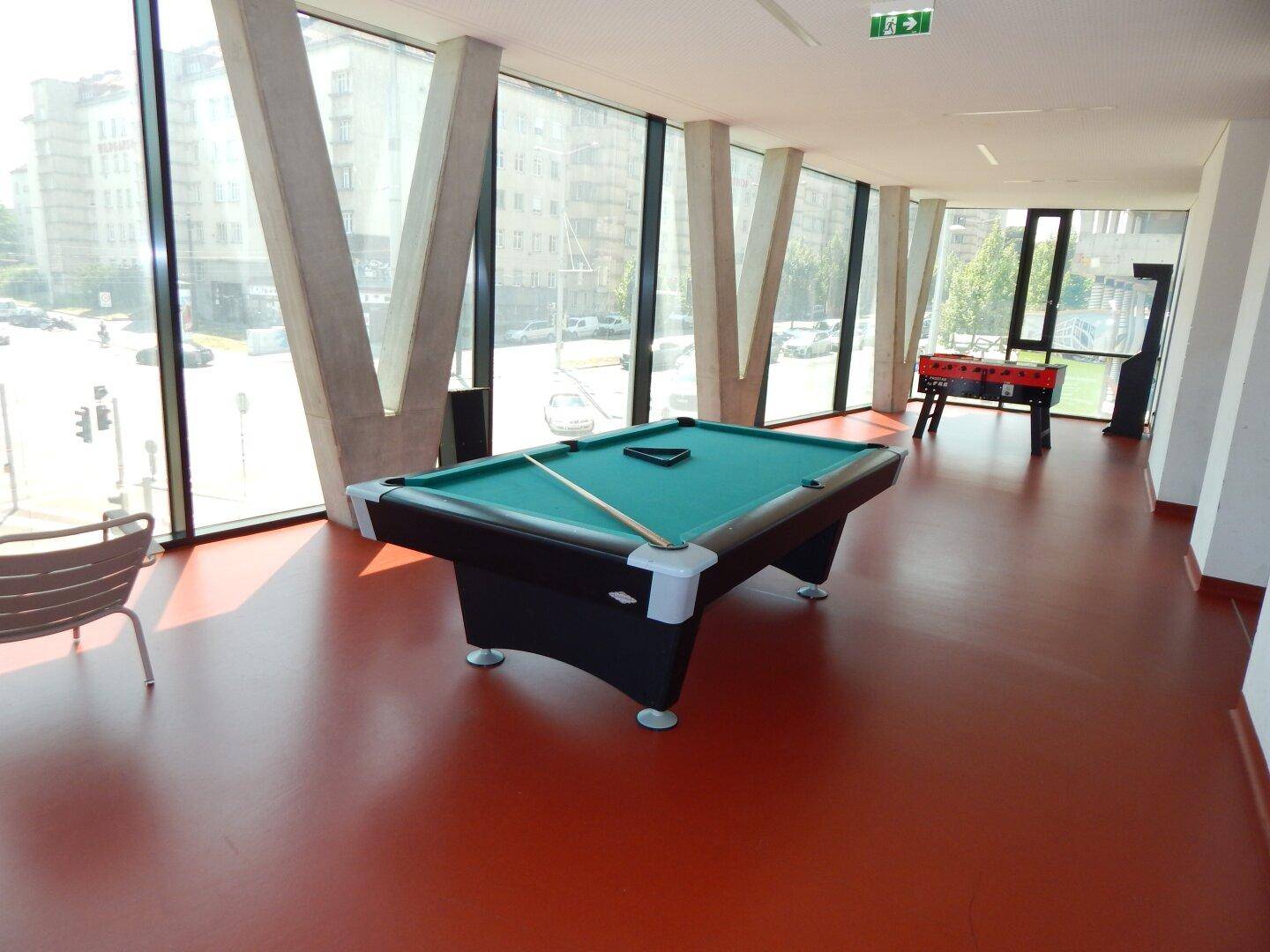 Spielzimmer mit Billardtisch und Tischfußball, umgeben von großen Fenstern.