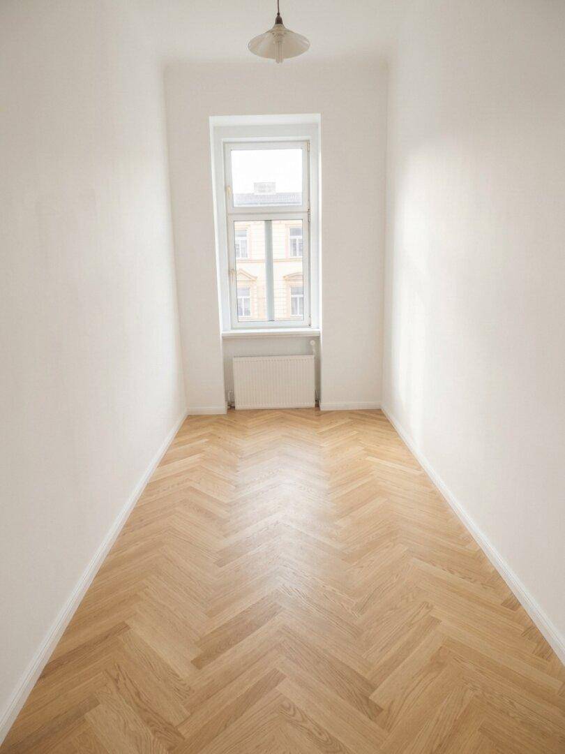 Schmaler Raum mit Fischgrätparkett und einem hohen Fenster, das Tageslicht spendet.