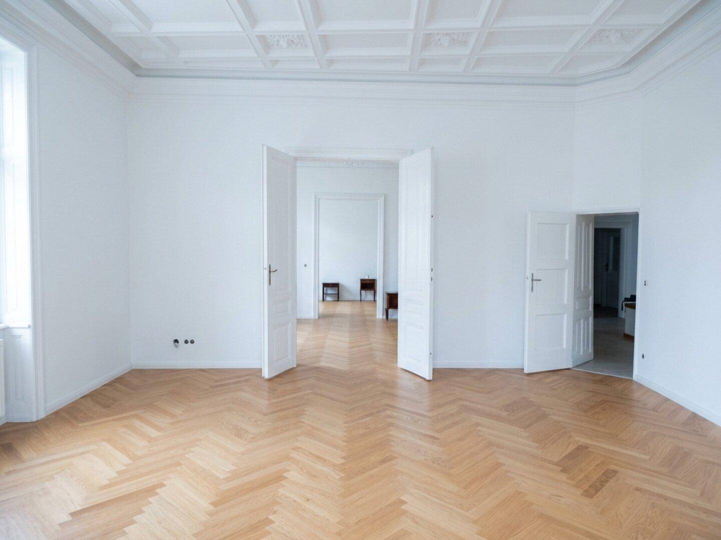 Geräumiges Wohnzimmer mit Fischgrätparkett und einer eleganten Kassettendecke, die den Altbaucharakter unterstreicht.