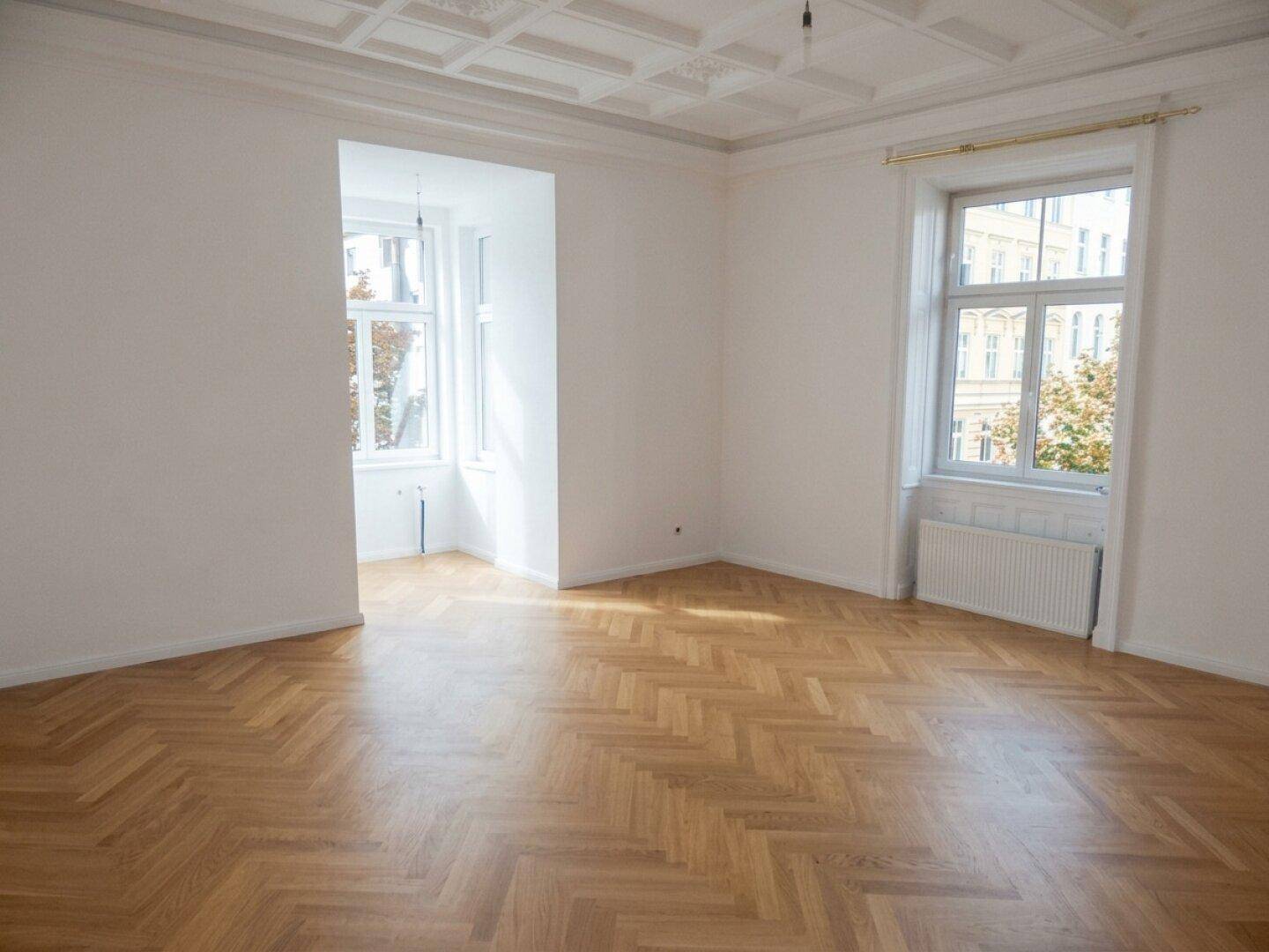 Großzügiges Zimmer mit Fischgrätparkett und zwei großen Fenstern, die den Raum hell erscheinen lassen.