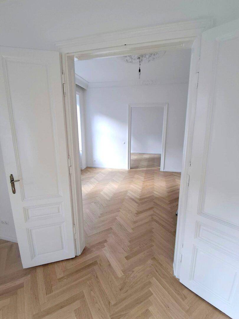 Blick durch hohe Flügeltüren in einen weiteren hellen Raum mit Fischgrätparkett und hohen Decken.