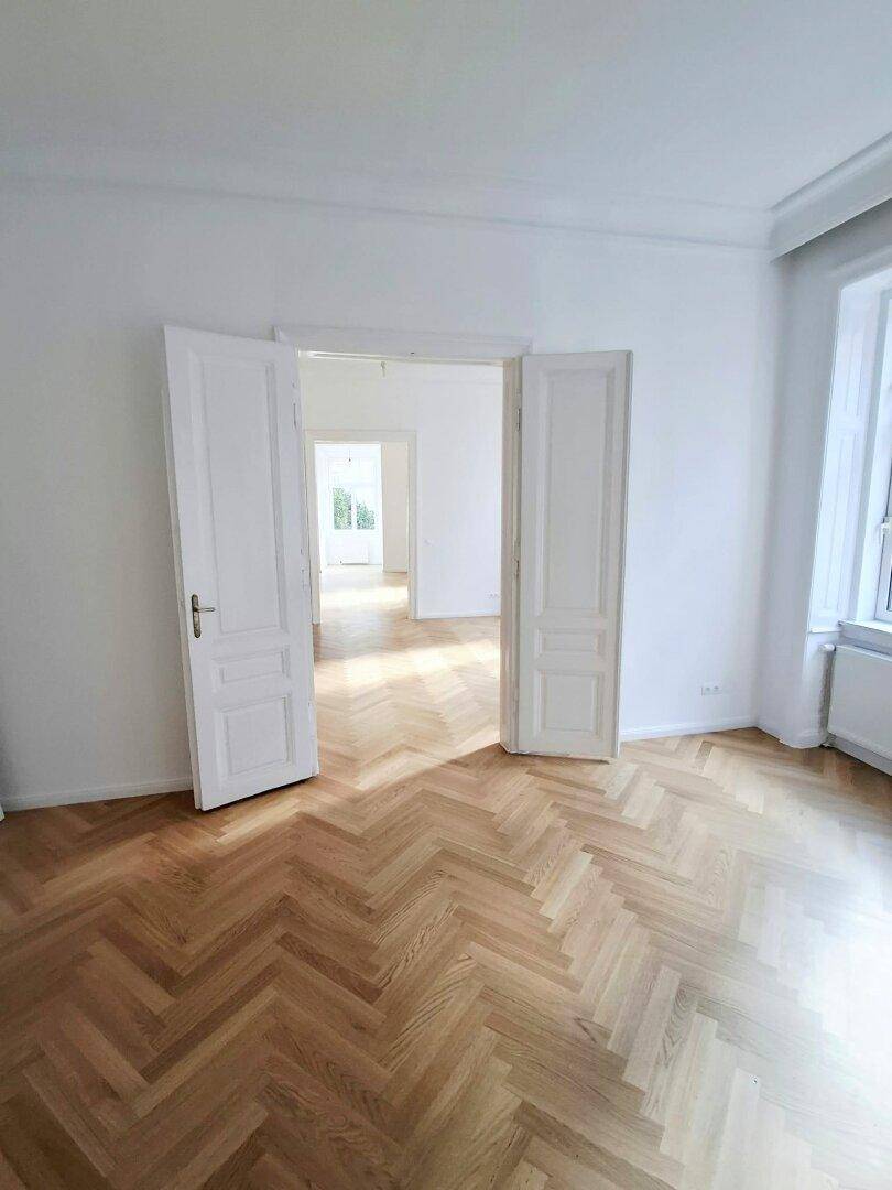 Heller Raum mit Fischgrätparkett und hohen Flügeltüren, die eine offene Raumgestaltung ermöglichen.