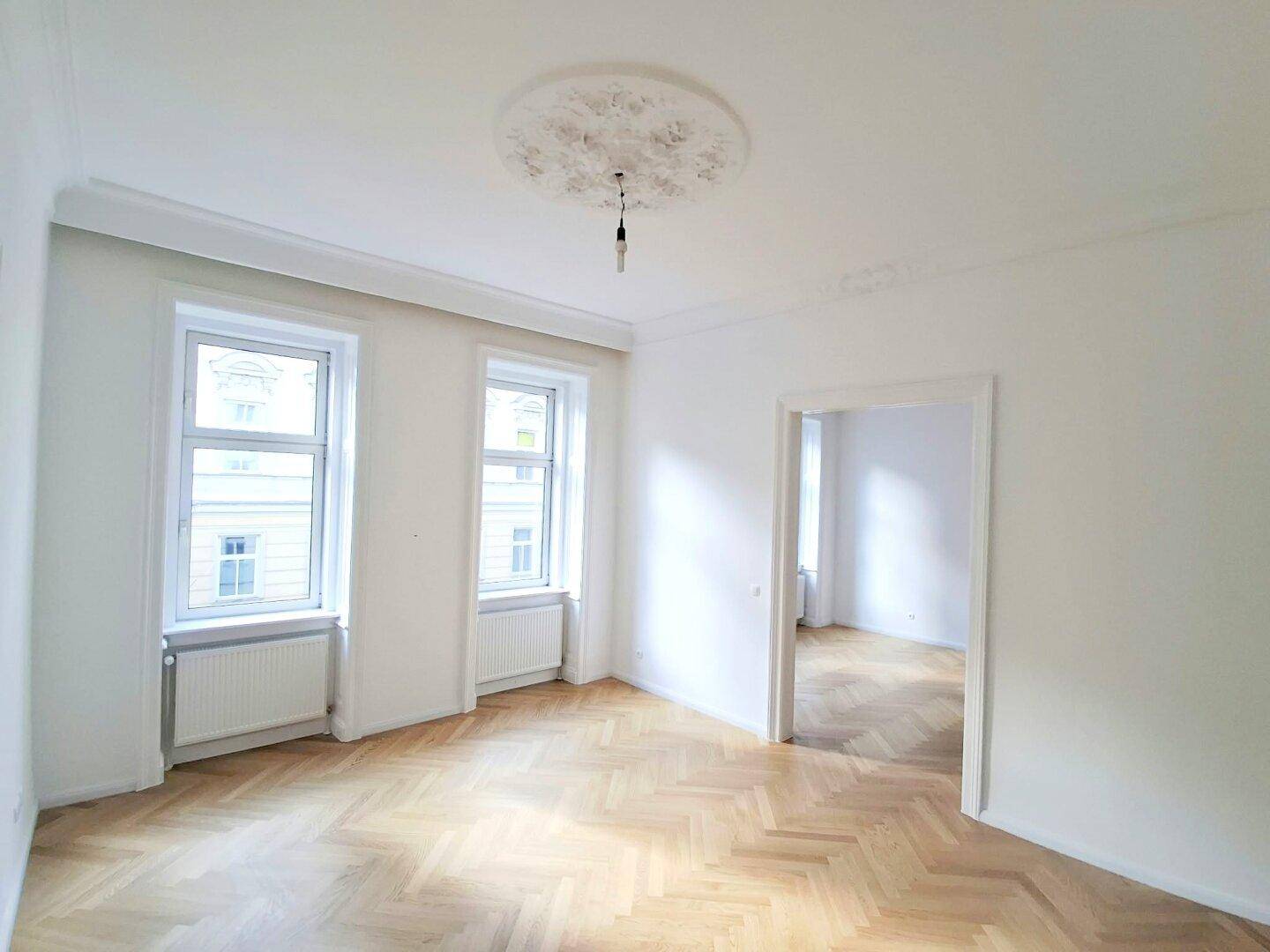Geräumiges Zimmer mit Fischgrätparkett, hohen Decken und einem dekorativen Deckenmedaillon.