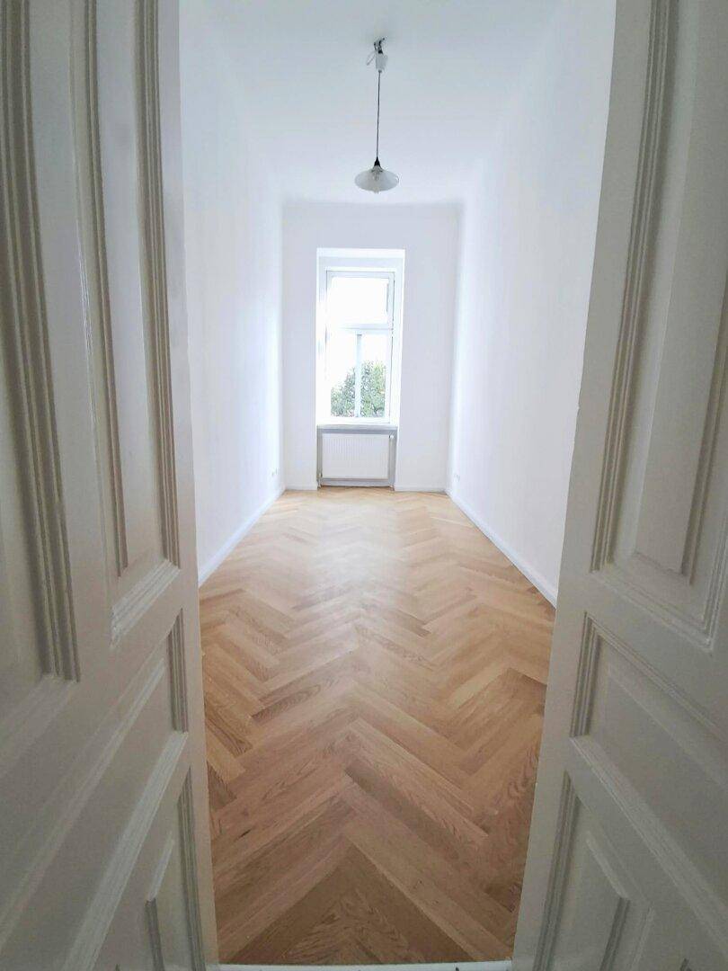 Langer, heller Raum mit Fischgrätparkett und einem Fenster am Ende, das Tageslicht spendet.