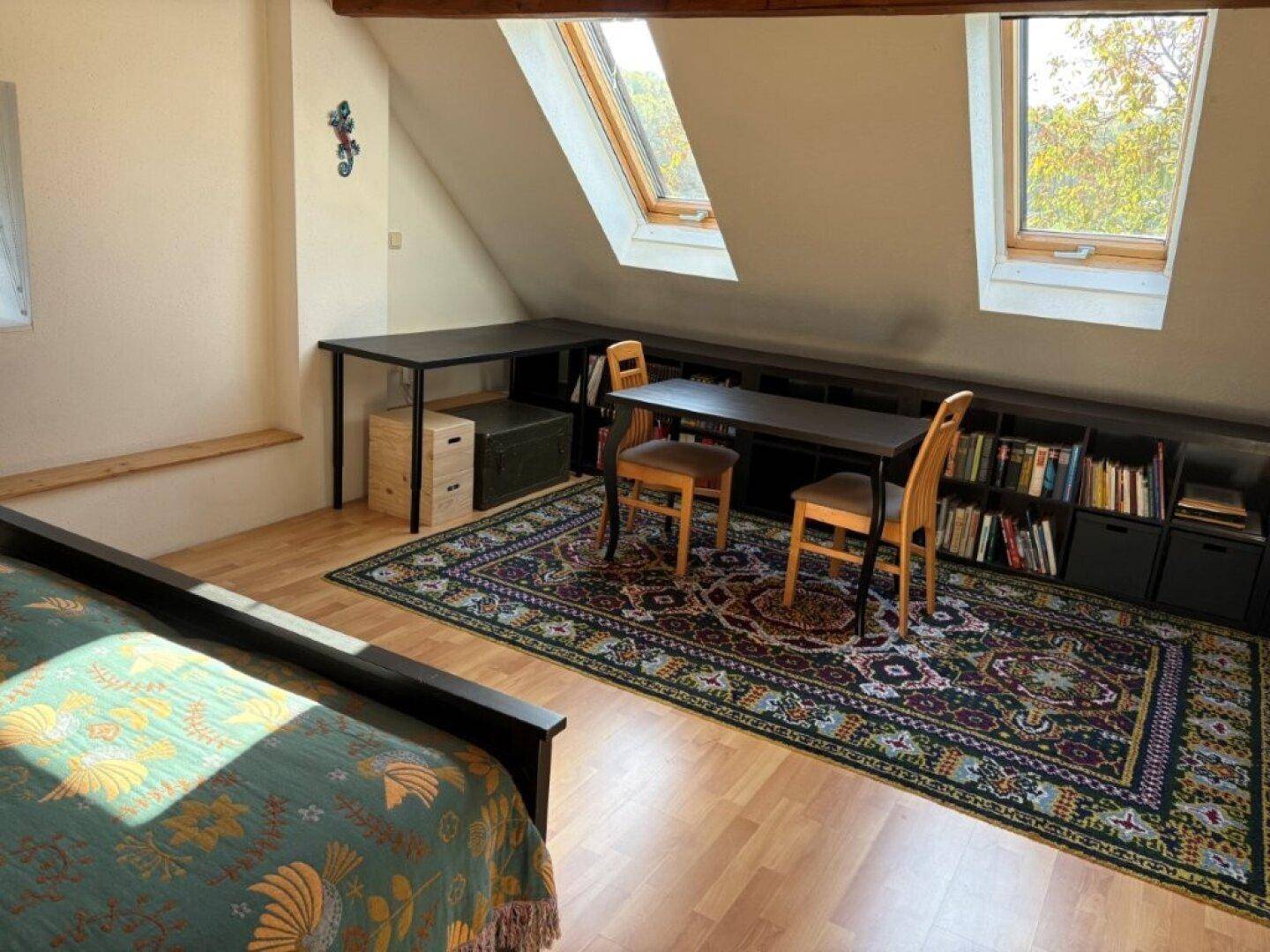 Dachgeschosszimmer mit Holzbalken, Dachfenstern, Bett und Arbeitsbereich mit Bücherregal.