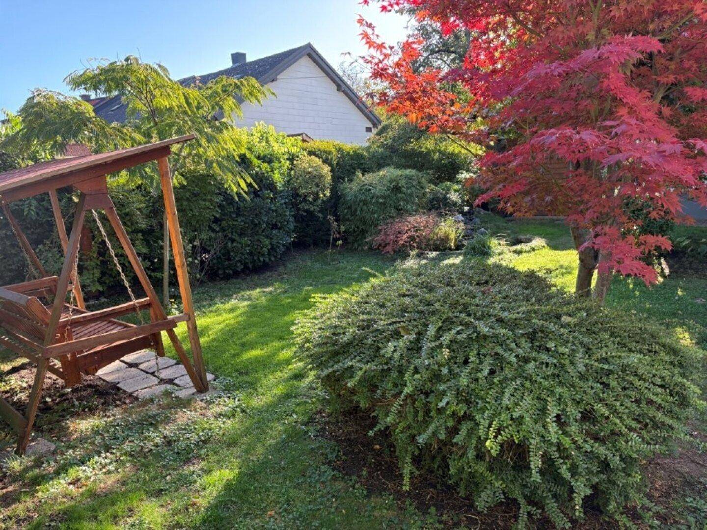 Sonniger Gartenbereich mit Holzschaukel, üppiger Bepflanzung und Herbstlaubfärbung.