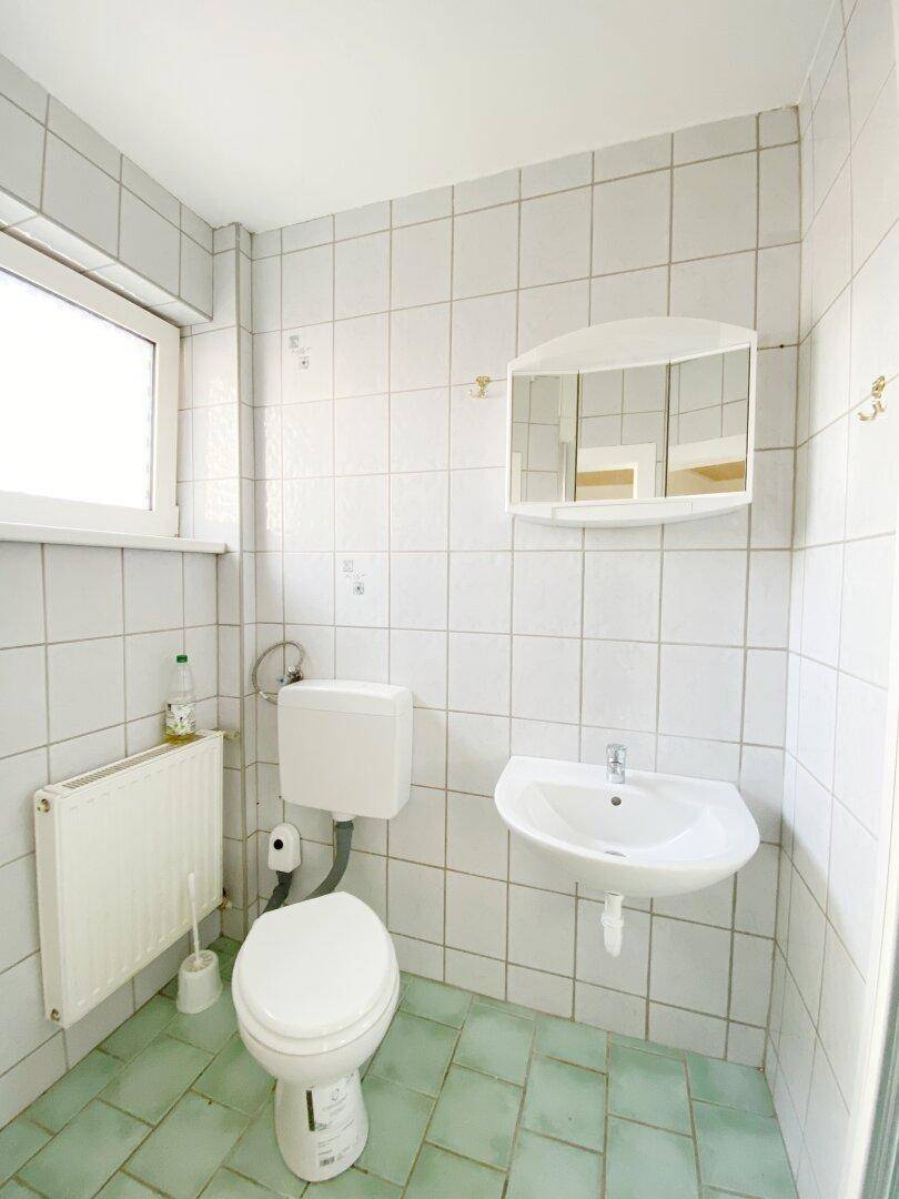 Badezimmer mit WC, Waschbecken, Heizkörper und Fenster, ausgestattet mit weißen und hellgrünen Fliesen.